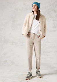 Luddig krämfärgad jacka med knappar, vit t-shirt, ljusbeige byxor och sneakers med grön ton, toppad med en blå stickad mössa.