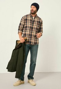 Karierte Flanellhemd in Beige, Braun und Marineblau; Jeans; hält eine dunkelgrüne Jacke; beige Turnschuhe; trägt eine marineblaue Mütze.