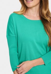 Žalias megztinis su apvalia iškirpte ir trigubomis rankovėmis. Yra raukšlėtos rankogaliai ir minkšta tekstūra.