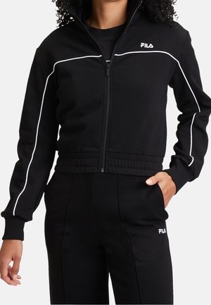 Kobieta ubrana w czarny dres FILA z białym obszyciem, kurtka zapinana na zamek, elastyczny pas, ręka w kieszeni czarnych spodni.
