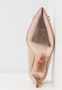 Escarpin beige à talon aiguille avec une tige lisse et texturée, un bout pointu, et une semelle métallique rose doré brillante ornée de motifs côtelés et d'un détail logo.