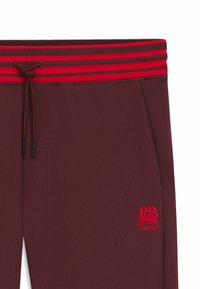 Burgundy joggers with a red striped elastic waistband, featuring a drawstring, side pocket, and embroidered logo detailing.