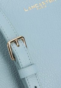 Borsa in pelle blu chiaro con una finitura testurizzata, caratterizzata da una fibbia dorata e dettagli di cucitura. Nome del marchio "Lancaster Paris" impresso in oro.