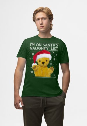 Grøn t-shirt med en gul bamse iført en rød nissehat. Teksten siger "JEG ER PÅ SANTAS UHYGGELIGE LISTE," omgivet af hvide snefnug.