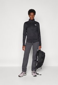 Črna zip-up jakna, sivi hlače in svetlo vijolični pohodniški čevlji. Drži črn nahrbtnik, ki vsebuje zmogljive zunanje materiale in eleganten design.
