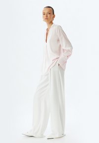 Blusa di seta rosa chiaro con scollo a V, maniche lunghe e polsini abbottonati, abbinata a pantaloni bianchi a gamba larga e scarpe a punta.