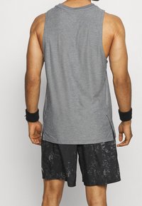 Šedý bezrukávový sportovní top s bočními rozparky, kombinovaný s černými šortkami s potiskem a černým náramkem, ukazuje sportovní design.