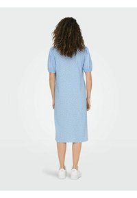 Sininen puolipitkä mekko lyhyillä pörrösateilla, jossa on hienovarainen kuvio. Malli on pukeutunut valkoisiin lenkkitossuihin. Takana oleva näkymä on esitetty.