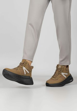 Osoba nosząca jasnoszare spodnie i brązowe wysokie trampki z czarnymi podeszwami i białymi bocznymi paskami, jedna stopa lekko uniesiona.