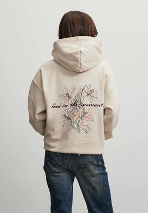 Person iført beige hættetrøje med blomsterdesign og teksten "live in the moment" på ryggen, kombineret med blå jeans, vendt væk fra kameraet.