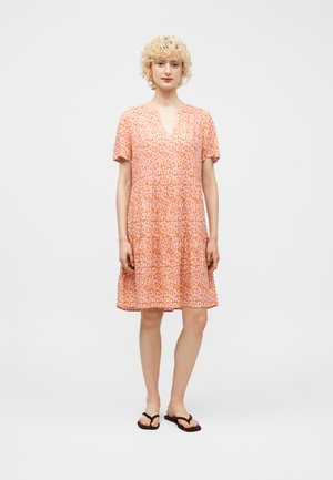 Kobieta o kręconych blond włosach, ubrana w pomarańczowo-różową sukienkę w krótkim rękawie z wzorem i czarne klapki, stojąca na tle jednolitego tła.