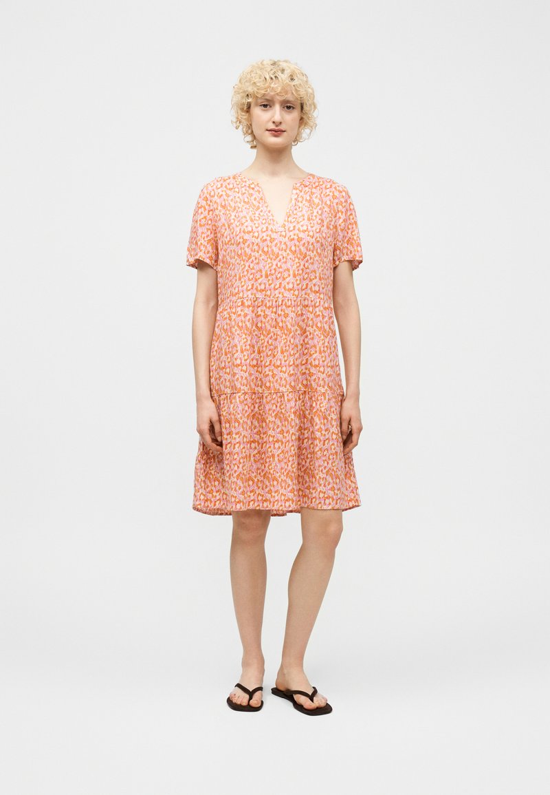 Γυναίκα με ξανθά, σγουρά μαλλιά, που φοράει ένα κοντομάνικο φόρεμα με πορτοκαλί και ροζ μοτίβα και μαύρες σαγιονάρες, στέκεται μπροστά σε ένα μονόχρωμο φόντο.