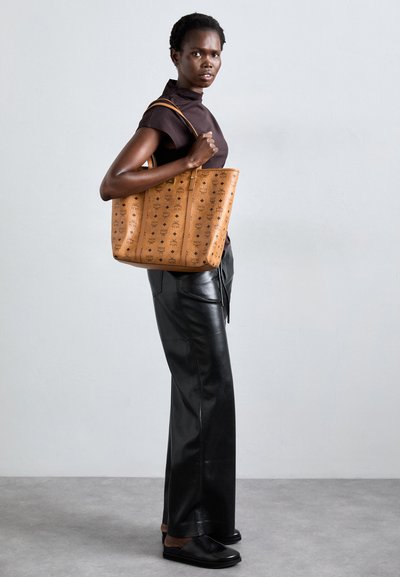 Woman wearing black top and leather pants holds large tan patterned tote bag standing against plain gray wall.