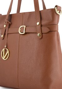 Sac fourre-tout en cuir marron avec une finition texturée, accents en métal doré et un pendentif rond affichant le nom de la marque. Deux poignées robustes.