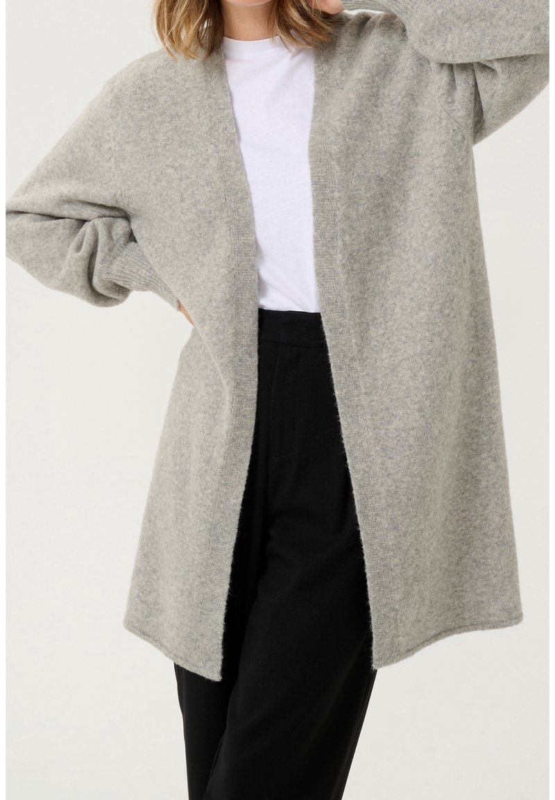 Femme portant un cardigan gris clair en laine à ouverture frontale, un t-shirt blanc à col rond et un pantalon noir taille haute, posant avec la main sur la hanche.