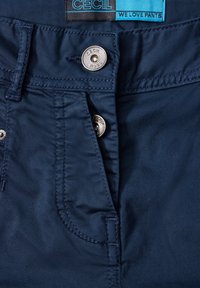 Mörkblå byxor med en mjuk textur, med en metallknäppning, sömnadsdetaljer och en etikett som säger "CECIL WE LOVE PANTS."