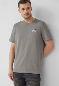 Szara bawełniana koszulka z okrągłym dekoltem, krótkimi rękawami i małym czarnym logo graficznym na piersi. Noszona z ciemnymi dżinsami.