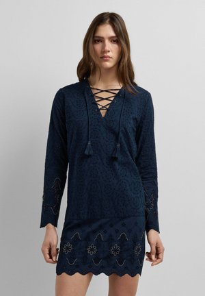 Vrouw draagt een marineblauwe jurk met lange mouwen, een vetersluiting aan de halslijn en een geborduurde bloemenrand met een geschulpte zoom, staand voor een effen grijze achtergrond.