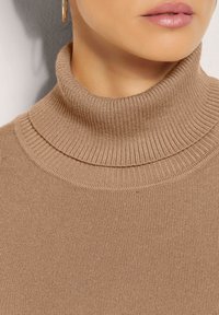 Taupe Strick-Turtleneck-Pullover mit gerippter Textur, figurbetontem Schnitt und hohem Kragen. Der Stoff wirkt weich und leicht.