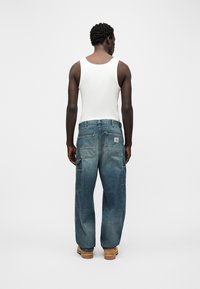 Άνδρας με άσπρο φανελάκι, φαρδιά ξεβαμμένα μπλε τζιν με πολλές τσέπες και καφέ εργασιακές μπότες, στέκεται με την πλάτη προς το μέρος της κάμερας σε απλό γκρι φόντο.