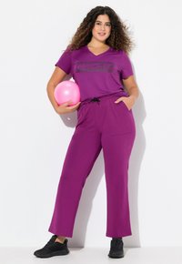 Sportovní fialový outfit zahrnující tričko s výstřihem do V a grafickým designem a volné kalhoty s kapsami. Drží se růžový cvičební míč.