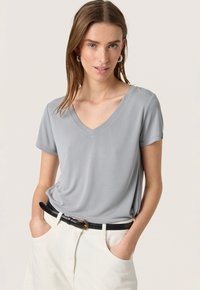 Szary t-shirt z dekoltem w serek, wykonany z miękkiego materiału; krótkie rękawy, luźny krój, noszony z białymi spodniami o wysokim stanie i czarnym paskiem.