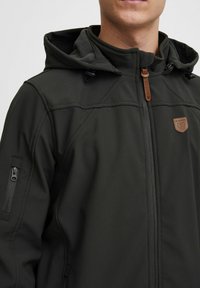 Mørkegrøn jakke med lynlås, justerbar hætte og struktureret stof med en lille logo-patch og lynlåslomme på ærmet.
