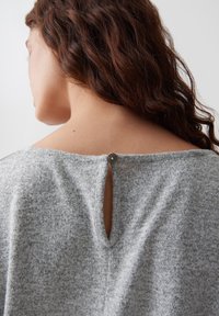 Femme avec des cheveux bruns ondulés portant un haut en maille gris boutonné dans le haut du dos, montrant une petite ouverture en forme de trou de serrure sous le bouton.