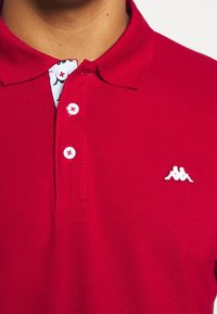 Polo rouge avec une finition texturée, doté d'un logo brodé blanc et d'une bordure contrastante à motif au col. Patte de boutonnage à trois boutons.
