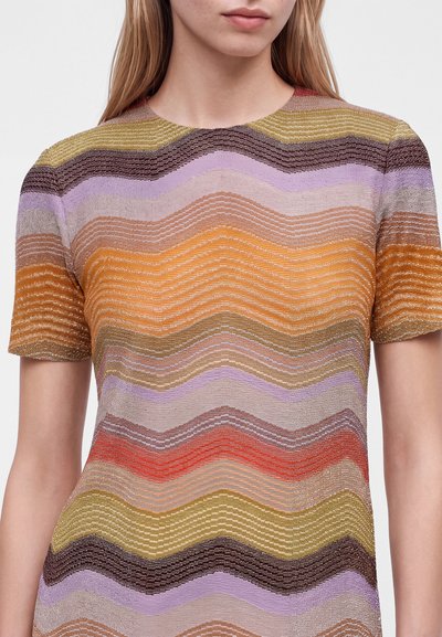 Femme portant un top en maille à manches courtes avec des rayures ondulées horizontales de couleur jaune, orange, violet, rouge, marron et beige.