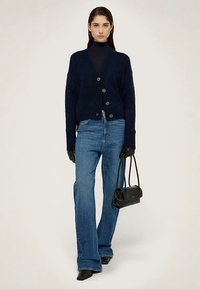 Tmavě modrý pletený kardigan s velkými knoflíky, spojený s modrými rozšířenými džíny s vysokým pasem. Doplňky zahrnují černé rukavice a malou kabelku.