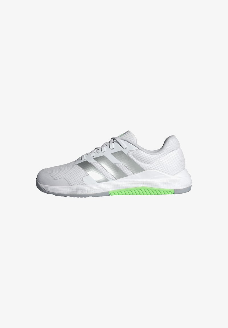 Chaussure de sport blanche avec des bandes argentées, une tige en mesh, une semelle blanche et une section de grip vert texturé sur le bord extérieur de la semelle.