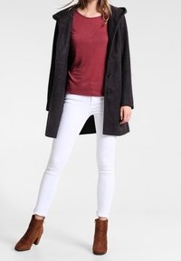Donna che indossa un cappotto con cappuccio grigio scuro, un top bordeaux, jeans skinny bianchi e stivaletti alla caviglia in camoscio marrone, in piedi contro uno sfondo semplice.