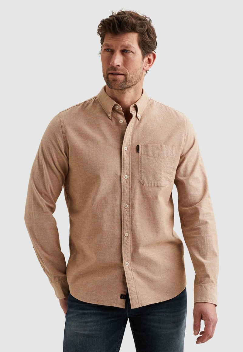 Homme portant une chemise beige à boutons avec poche poitrine et un jean foncé, regardant vers la gauche sur un fond uni.