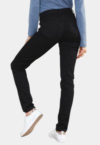Schwarze Skinny-Jeans mit hohem Taillenbund, aus einem glatten Material und mit zwei Gesäßtaschen. Kombiniert mit weißen Sneakers und einem blauen Oberteil.