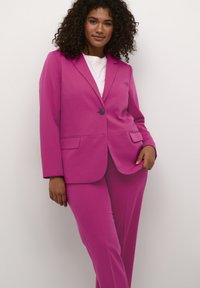 Abito rosa composto da un blazer sartoriale con un solo bottone e pantaloni. Tessuto liscio con due tasche frontali sul blazer. Camicia bianca sotto.