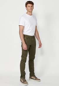 Pantalones de mezclilla slim-fit en verde oliva con cinco bolsillos y presillas tradicionales para el cinturón, combinados con una camiseta blanca básica y zapatillas marrones con estampado.