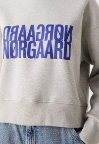 Personne portant un sweat gris avec un texte bleu en miroir et droit "NORGAARD" sur un jean bleu, main droite dans la poche.