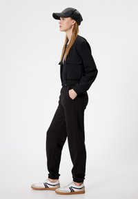Zwart cropped bomberjack met twee voorzakken, zwarte joggers en witte sneakers met zwarte accenten. Model draagt een zwarte pet.