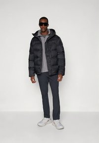 Svart dunjakke med hette, grå hettegenser under, mørke bukser og hvite joggesko. Modellen har på seg mørke solbriller. Jevn tekstur.