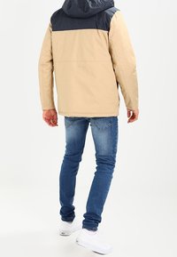Chaqueta con capucha en color beige y azul marino, con frente con cremallera, sección superior en contraste y corte recto, combinada con jeans azules y zapatillas blancas.