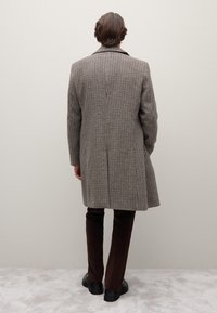 Geruite wollen jas in bruin en crème, tot op de knieën met een enkele inkeping en reverkragen, gecombineerd met donkerbruine ribfluwelen broek en zwarte schoenen.