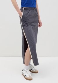 Grijze maxi-rok met een hoge zijsplit en elastische tailleband, voorzien van een contrasterende crème zijstreep; gedragen met witte sneakers.