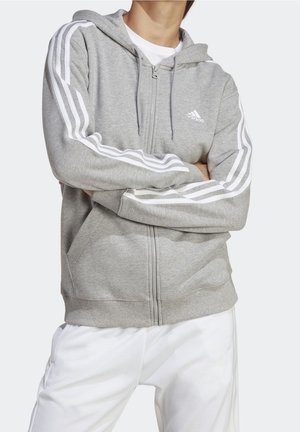Osoba nosící šedou mikinu Adidas na zip s bílými pruhy na rukávech a bílé kalhoty, ruce překřížené přes hruď.