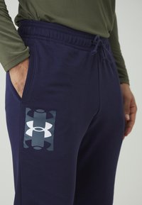 Navy bikses ar uzrakstu, kurā redzams izteikts balts Under Armour logos kreisajā augšstilbā, izgatavotas no mīkstas, teksturētas auduma ar elastīgu jostasvietu.