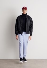 Μαύρο μπουφάν bomber πάνω από μαύρο πουλόβερ, σε συνδυασμό με ανοιχτόχρωμα μπλε φόρμες που έχουν κείμενο και μαύρα αθλητικά παπούτσια. Κόκκινο καπέλο στο κεφάλι.