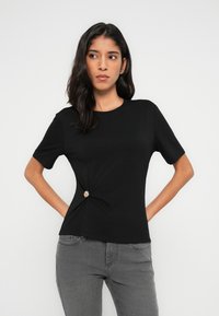 Μαύρο κοντομάνικο μπλουζάκι από μαλακό ύφασμα με στρογγυλή λαιμόκοψη και κόμπο στη μέση, κλειστό με χρυσό κουμπί.