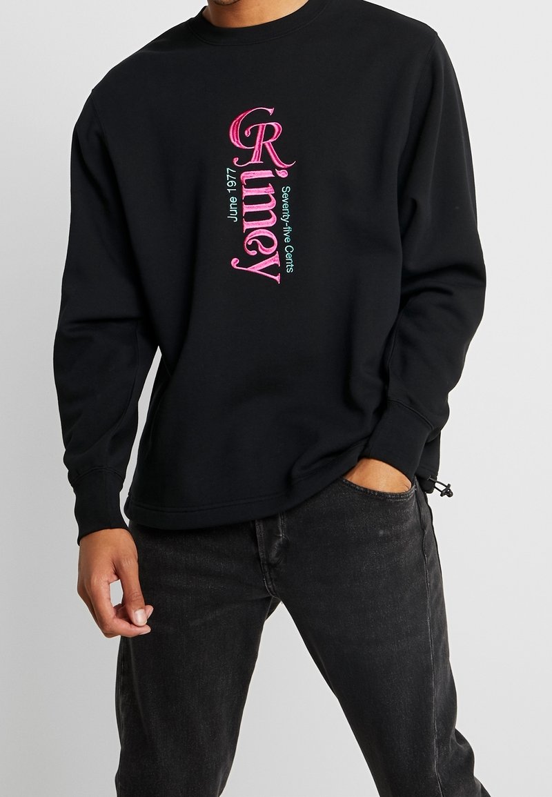 Uomo che indossa un maglione nero con testo verticale rosa e azzurro chiaro "Rimey Giugno 1977 Settantaquattro Cents" e jeans neri.