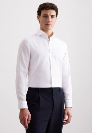 Uomo che indossa una camicia bianca a maniche lunghe con bottoni e pantaloni scuri blu navy, in piedi contro uno sfondo chiaro e semplice.
