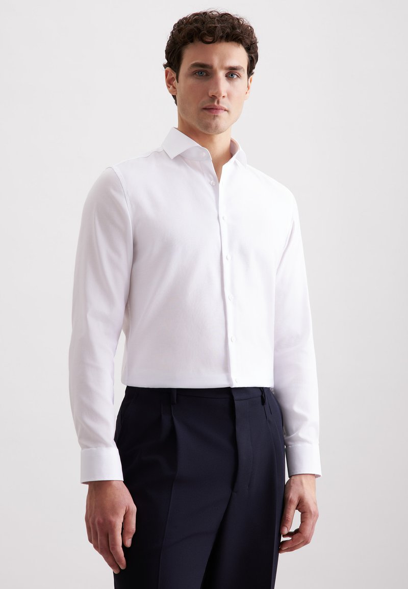 Uomo che indossa una camicia bianca a maniche lunghe con bottoni e pantaloni scuri blu navy, in piedi contro uno sfondo chiaro e semplice.